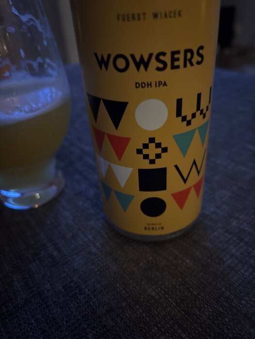 En burk Wowsers DDH IPA, färgglad med geometriska former, bredvid ett glas öl på ett bord.