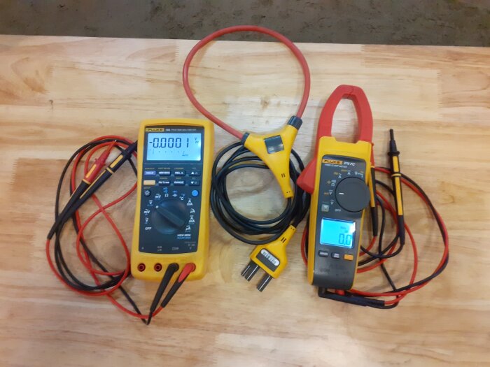 Två Fluke mätinstrument med kablar ligger på ett träbord, en digital multimeter och en tångamperemeter, redo för elektriska mätningar.