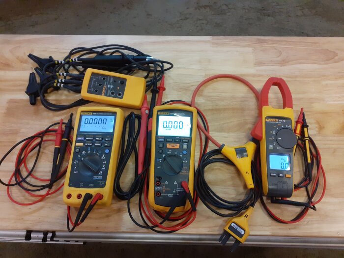 Tre digitala mätinstrument från Fluke på ett träbord, med kablar och mätspetsar, bland annat en tångamperemeter med trådlös funktion.