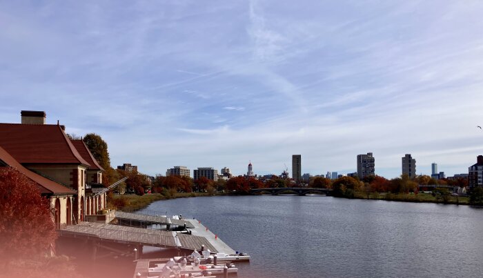 Flodlandskap med kaj, rödtakshus och stadssilhuett i bakgrunden med bro och röda höstträd.