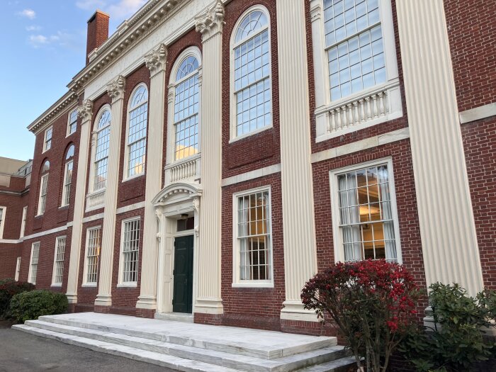 Röd tegelbyggnad med vita kolonner, stora fönster och grön entrédörr, omgiven av buskar och en vit stentrappa i förgrunden.