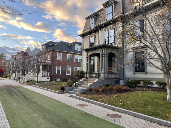 Gata med äldre hus i trä och tegel, omgiven av träd och stigande kvällssol på en molnig himmel.