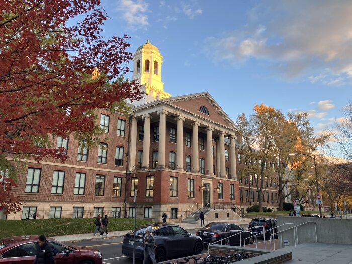 En tegelbyggnad med pelare och kupol, omgiven av höstfärgade träd. Människor och bilar på gatan framför byggnaden.