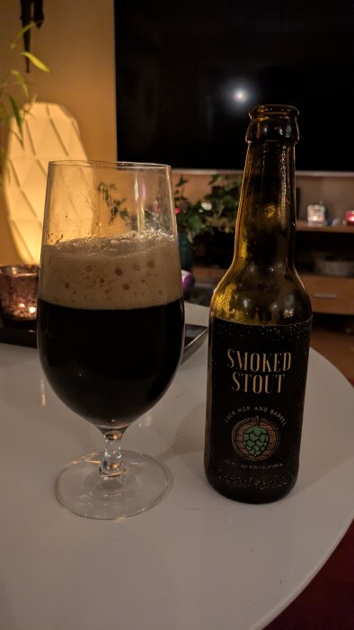 Ett glas med skummande mörk stout och en flaska märkt "Smoked Stout" står på ett bord i ett hemtrevligt rum.