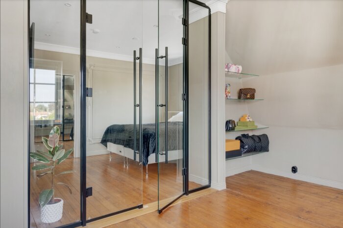 Spegelvägg i en walk-in-closet som visar en säng i master bedroom, med hyllor till höger och ett trägolv.