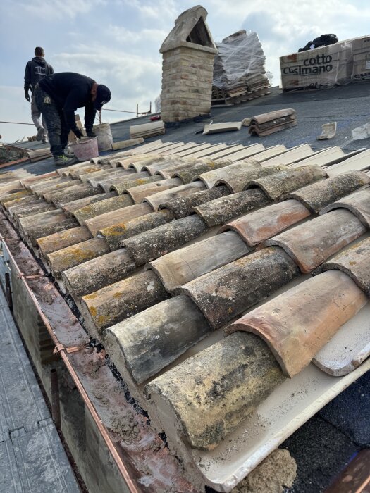 Två personer lägger taktegel på ett tak med nya pannor i botten och återbrukade ovanpå, intill en tegelstensskorsten.