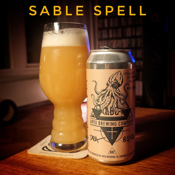 Glas med ljust öl och en burk från Apex Brewing Company med en bläckfiskillustration på en träyta, text "Sable Spell" syns ovanför.