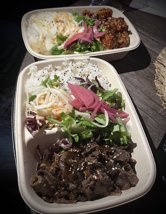 Två takeout-matlådor med japanska rätter, inklusive ris, marinerat kött, picklad rödlök och sallad på ett träbord.