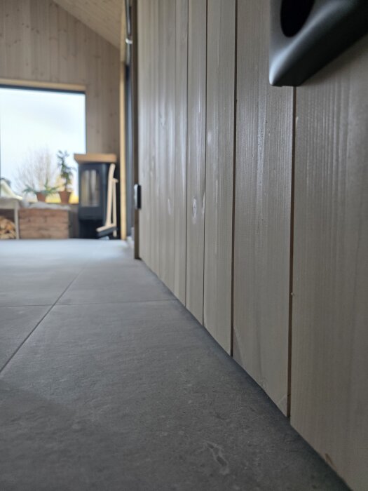 Närbild av en träpanelvägg med golvet nedanför, samt ett rum med utsikt mot fönster och vedspis i bakgrunden.