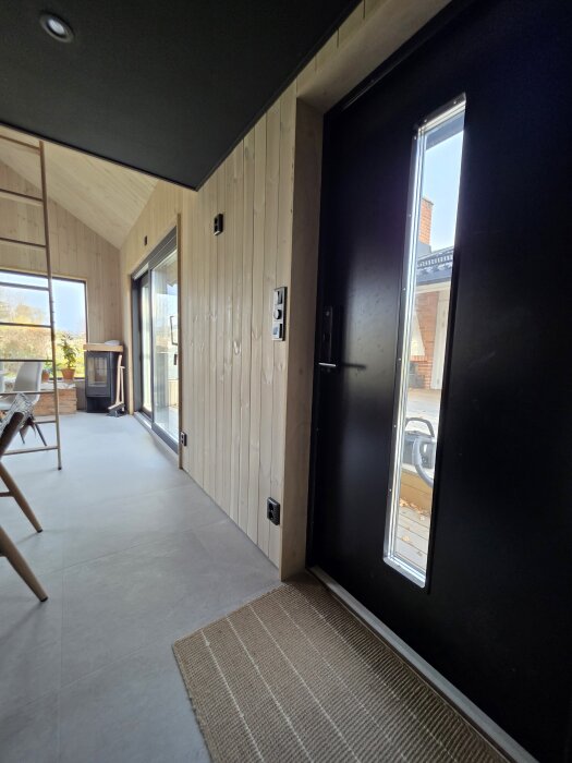 Modernt rum med svart tak och väggpanel, stor glasdörr, utsikt utåt. Samma färg på tak och vägg, lystergolv, inredning i trä.