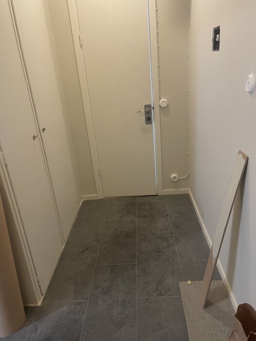 En hall med mörk klinker och ljusa väggar, en ljusgrå provplatta mot väggen.