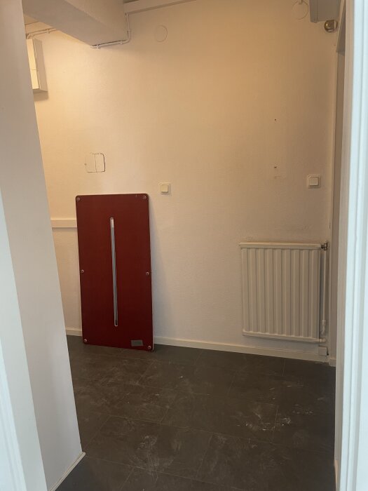 En hall med mörkt klinkergolv, vit vägg och ett rött objekt lutande mot väggen. En radiator syns på höger sida.