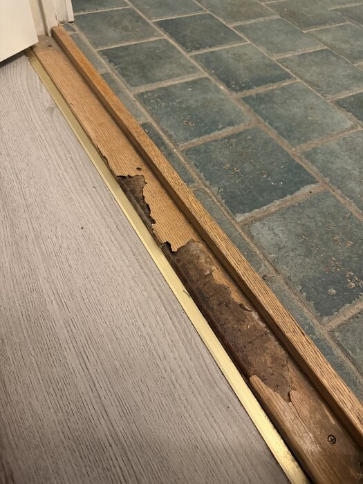 Sliten trätröskel mellan hall och toalett med synliga skador och metallist mot kaklad golvyta.