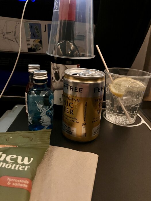 Dryckesutbud på ett flygplansbord med tonicvatten, små flaskor sprit, nötter och en glasdrink med citron.
