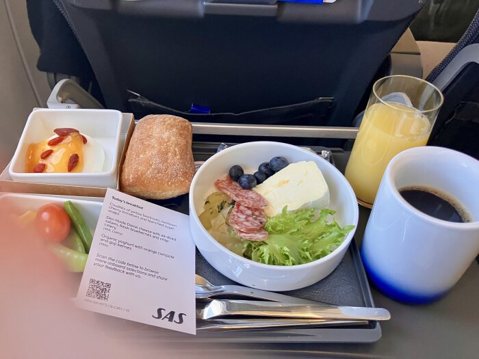 Frukost serverad på flygplan med yoghurt och bär, ciabattabröd, kallskuret, sallad, kaffe och apelsinjuice på bricka.