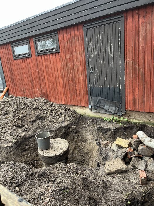 Grävd grop vid röd byggnad med synlig nedstigningsbrunn och jordhögar, intill en hög av stenar och tegel.