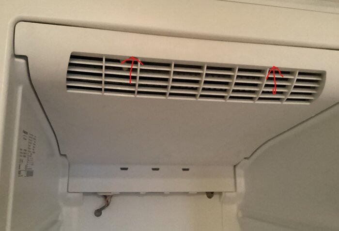 Ventilation på vit kylskåpsinsida med två röda pilar som markerar skruvhål i framkant.
