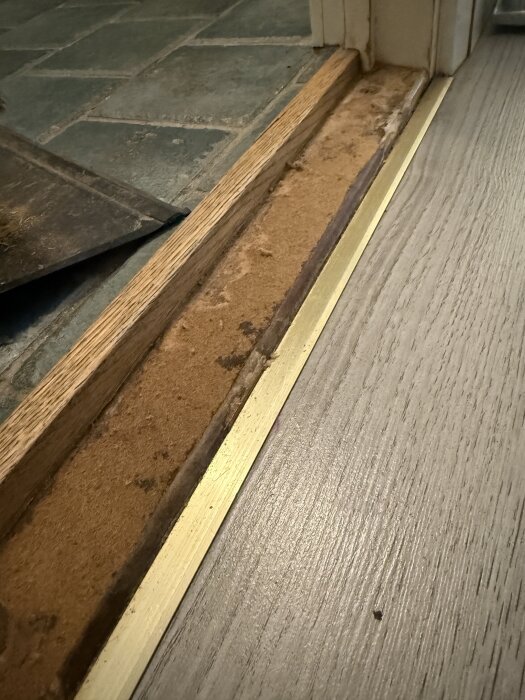 Tröskel mellan hall och toalett med borttagen yta, synlig trä- och metallist, samt skadat material, som visas efter ett renoveringsförsök.