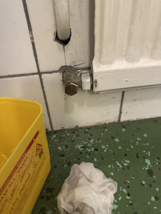 Vattenläcka vid radiatorventil på vitmålad vägg med grönt golv, gul plastbehållare och papperstrasa synliga i bild.