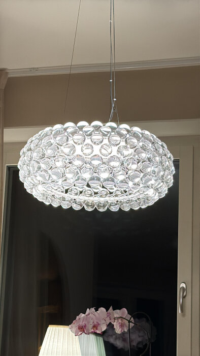 Modern kristallkrona med många glasprismor hänger i taket, ovanför en fönsterbräda med en bordslampa och blommande orkidé.