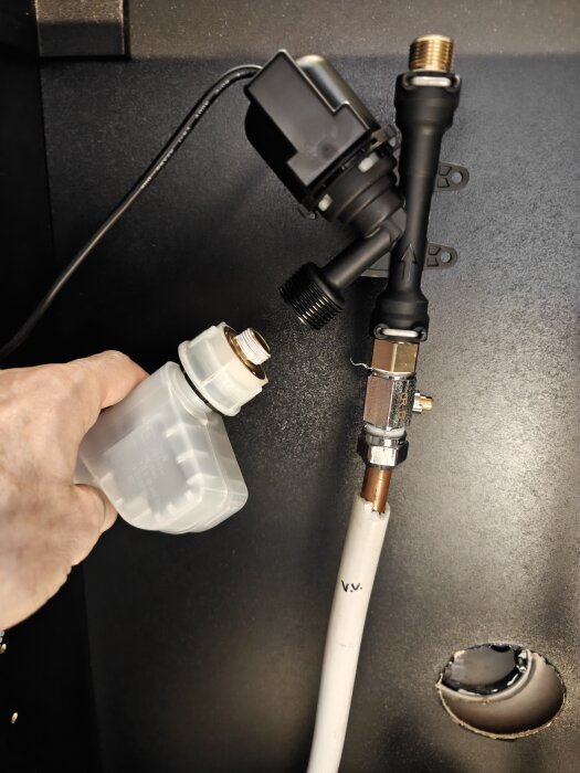 Hand som håller en plastdel nära en vattenanslutning med rör och kopplingar under diskbänk.