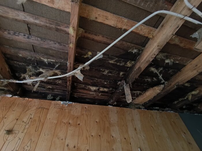 Garagetak med tydliga fuktskador och trasig isolering, samt exponerade träbalkar och kablar.