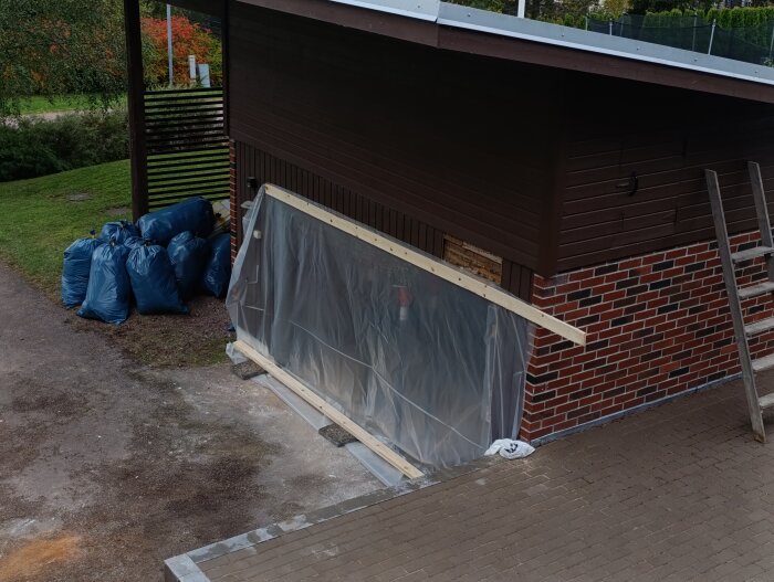 Garageport under renovering med plastöverdrag, byggmaterial och blå soppåsar bredvid. Trästege lutar mot garaget.