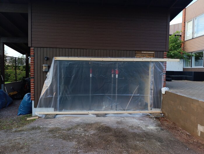 Garage under ombyggnad, ingång täckt med genomskinlig plast och trästöd. Bakom plasten syns delar av utbytta väggar och verktyg.