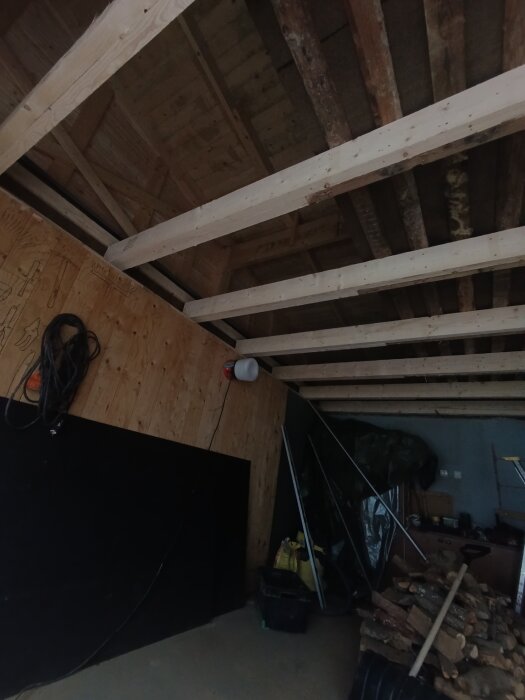Garagetakets innertak med nya träbjälkar och en hög ved i hörnet; vägbeskrivning för renovering med fuktskadad gips.