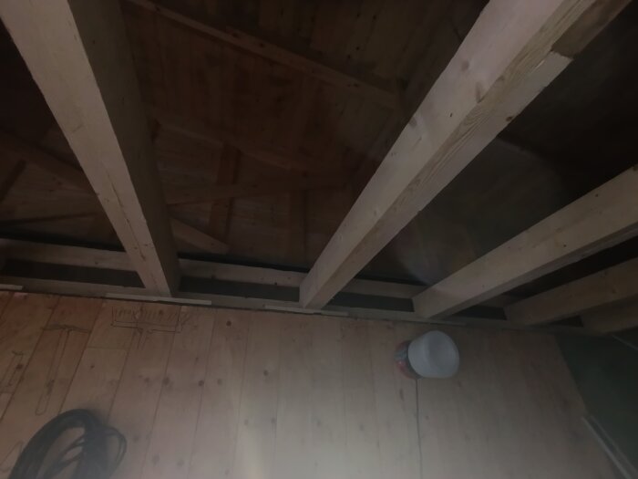 Takbjälkar av trä i ett garage samt några verktygsritningar på en trävägg. Sladd ligger på golvet, och en plastburk står lutad mot väggen.