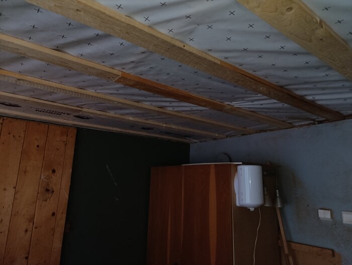 Garagets innertak med träbalkar och vit plastfolie delvis blottat, samt en del av en vägg med trädetaljer och ett skåp.