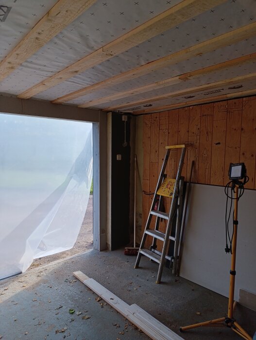Inuti ett modifierat garage med en bredare dörröppning, plasttäckt ingång, takunderarbete och en stege samt arbetslampa.