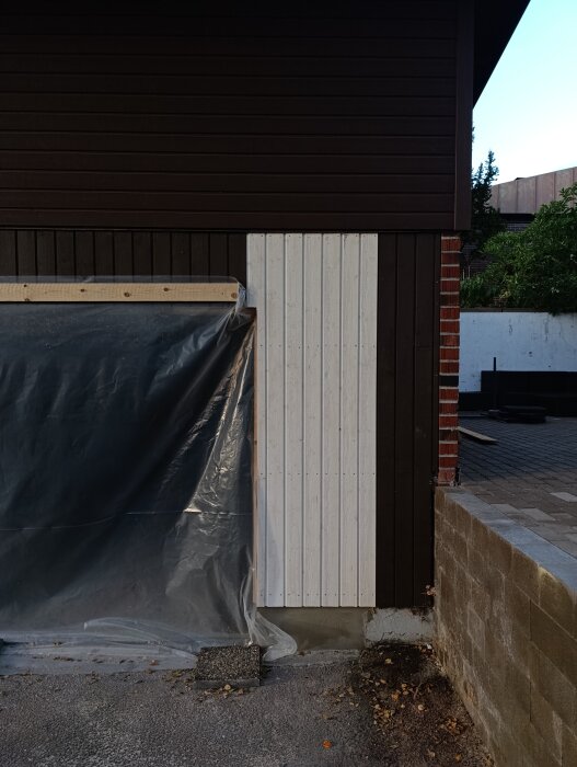 Breddad garageport med plastbit skyddar ingången. Träbeklädnad i olika färger, angränsande tegelvägg och en mur syns i bakgrunden.