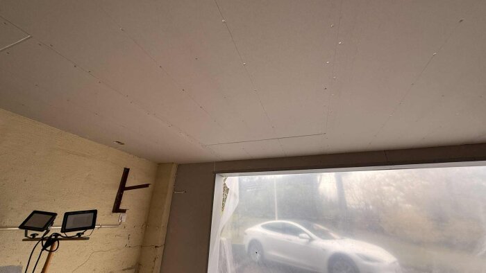 Garagetaket med ny gips, ett fönster och bil utanför. Arbetslampor syns mot en renoverad vägg.