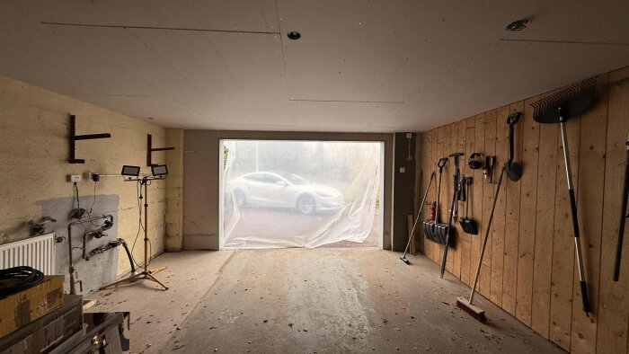 Garage med plastfilm över porten, verktyg hängande på väggen och en vit bil synlig utanför. Golv och tak visar tecken på renovering.