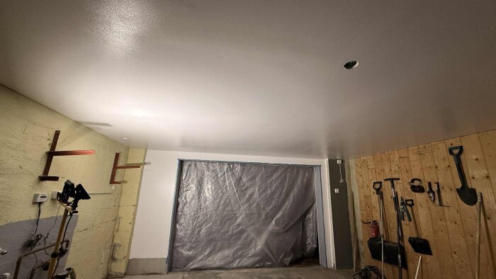 Renovering av garage med ett nyligen utökat dörrutrymme täckt av plast, samt synliga väggmonterade verktyg och ett nytt gipstak.