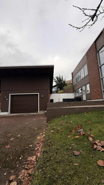 Modern fasad med brunt trä- och tegelhus, garageinfart och omgivande grönska, sett på en mulen dag.