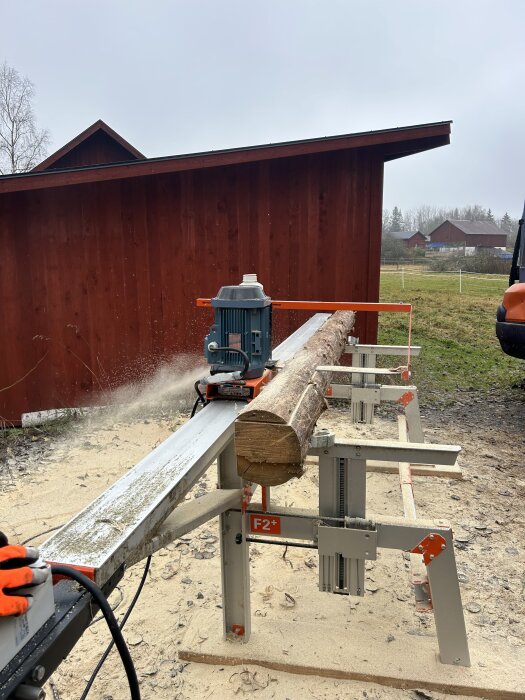 En sågmaskin bearbetar en trädstock utomhus framför en röd byggnad, med spån på marken.