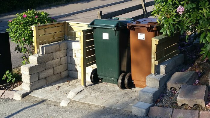 Två soptunnor placerade i en station byggd av trä och sten, med plats för ytterligare ett kärl. Rund murad yta syns intill växtlighet.
