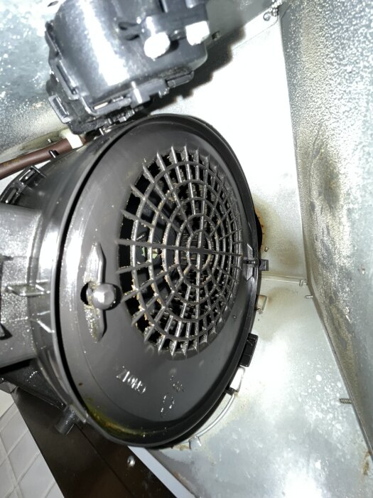 Närbild på insidan av en köksfläkt visar fläktens galler med piggar för montering, omgiven av metall och några synliga skruvar.