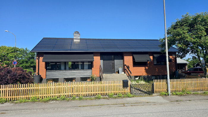 Tegelhus med soltak och svart dörr omgiven av trästaket, buskar och träd under klarblå himmel.