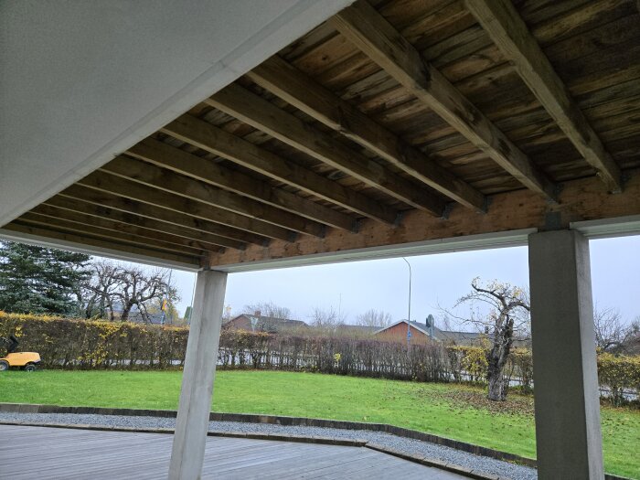Underifrån vy av en träkonstruktion med balkar och balkskor, ovanför en uteplats med grön gräsmatta och träd i bakgrunden.