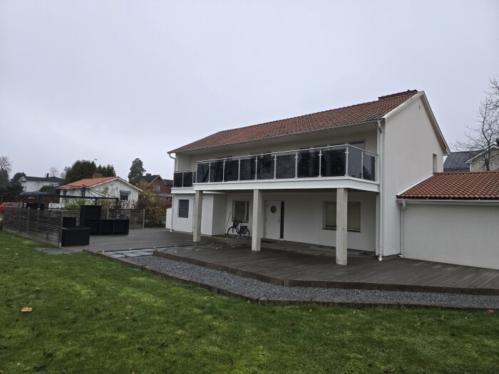 Vit tvåvåningsvilla med altan, balkong och röda tegeltak. Grön gräsmatta i förgrunden och cykel vid entrén. Grå himmel.