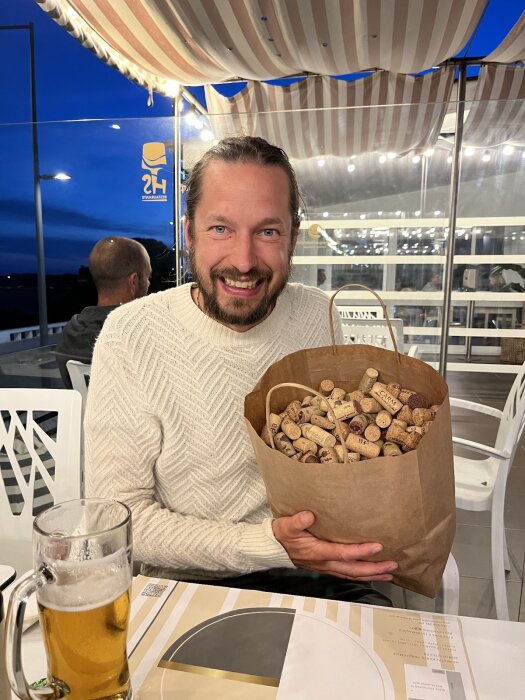 Man med långärmad tröja ler och håller en stor papperspåse fylld med vinkorkar. I förgrunden står ett glas öl på bordet.