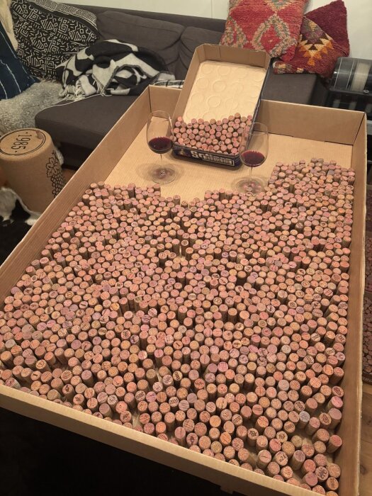 Kartong full med sorterade vinkorkar på ett bord, två vinglas med rödvin bredvid. Soffa och kuddar i bakgrunden.