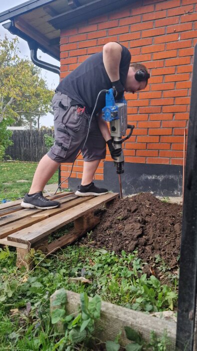 Person som använder en bilningsmaskin för att driva ner ett jordspett i marken framför en tegelvägg, stående på en lastpall.