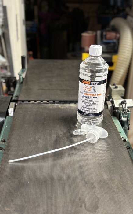 Flaska med CMT Formula 998 träsmörjmedel på en hyvelbänk i verkstad, med medföljande pump bredvid.