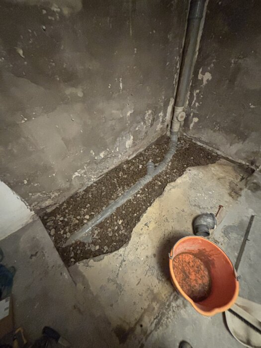 En nyligen lagad golvyta med avloppsrör synlig, omgiven av cement och grus. En orange hink står på sidan.