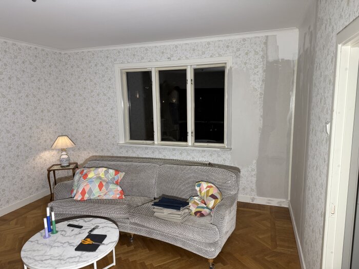 TV-rum under renovering med en blommig tapet, grå målad vägg, grå soffa med färgglada kuddar, runt soffbord och en bordslampa i hörnet.