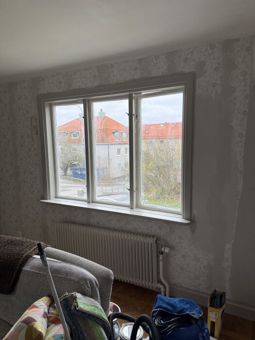 Fönster med utsikt mot byggnader, omgivna av mönstrade tapeter, och en soffa med filtar samt verktyg framför; del av renoveringsprojekt.